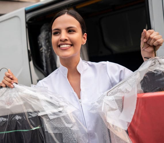 A smiling woman holds two plastic-covered garments on hangers while standing in front of a vehicle.