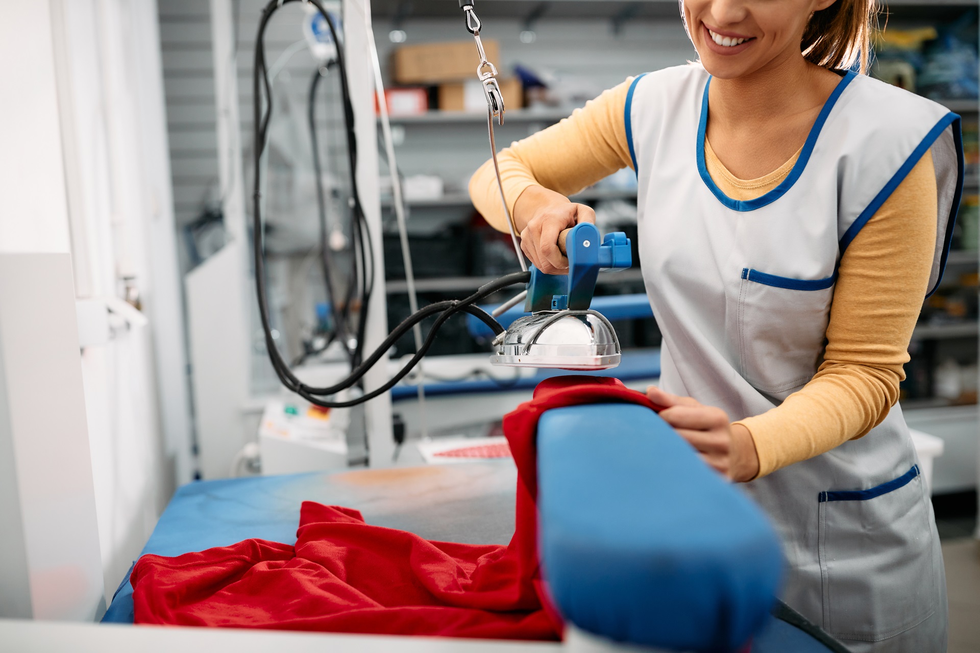 A person wearing a work uniform is smiling while ironing a red garment on a blue ironing board in a laundry facility.