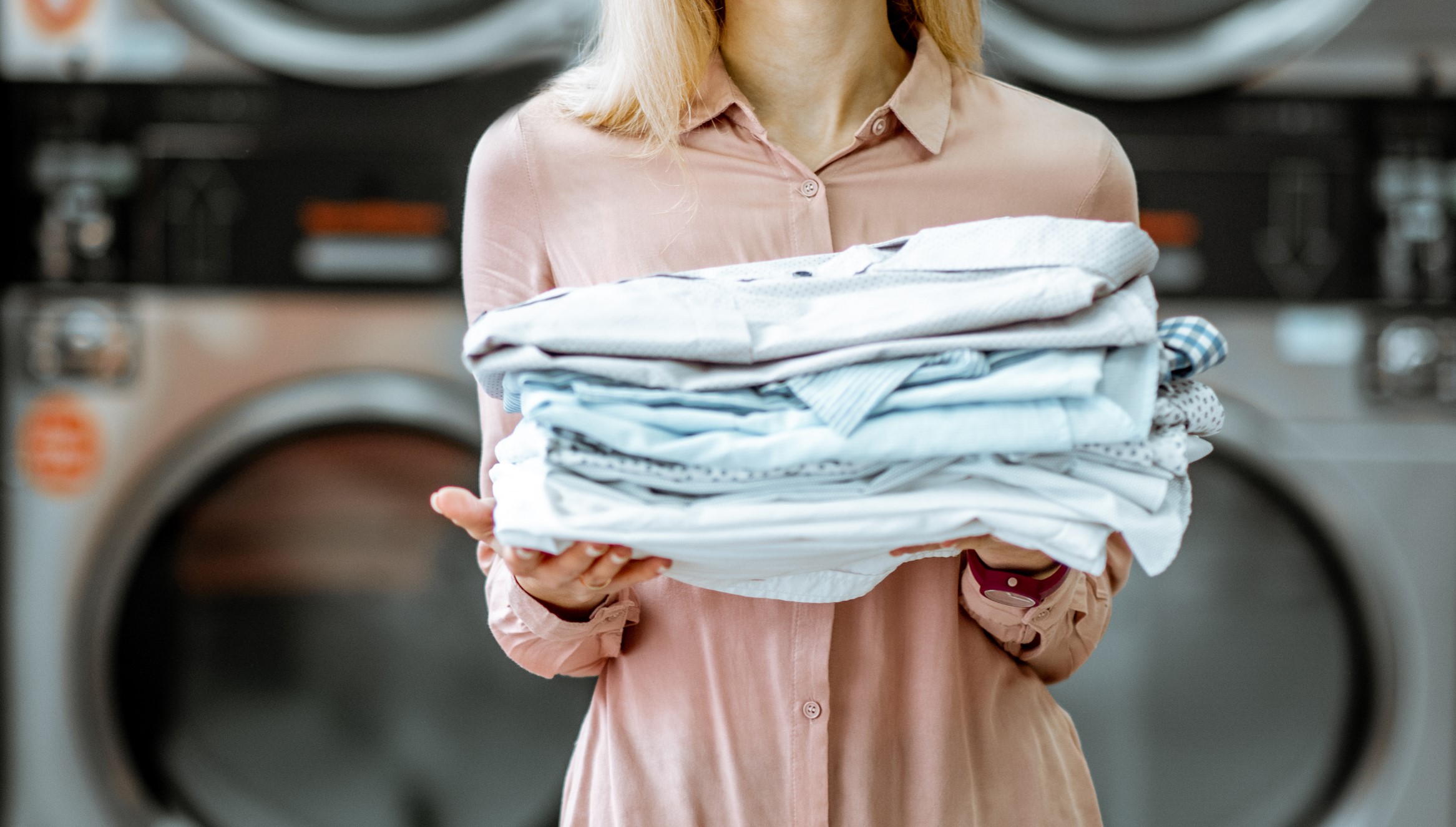 Person holding a stack of folded laundry in a laundromat with washing machines in the background.