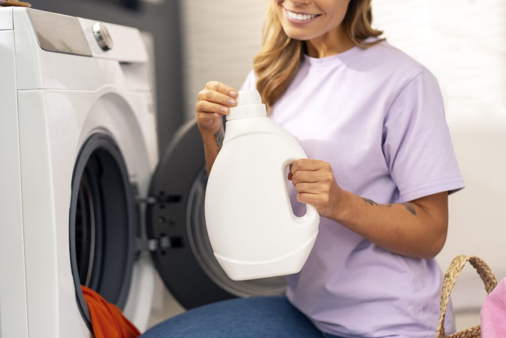 A person in a purple shirt holds a bottle of detergent, found at the best clothes cleaner near me, in front of an open washing machine filled with laundry.